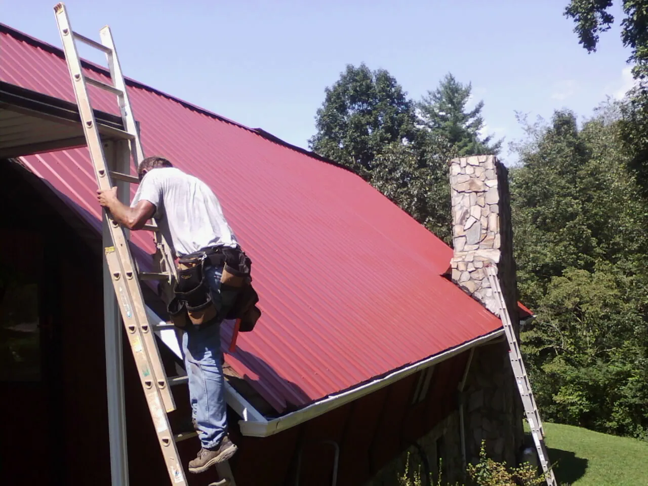 Roofer on a ladder beside a red metal roof and stone chimney — Soffit & Fascia Repair by Roofing Brothers in Mapleton Depot